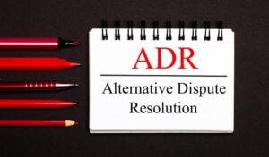 A white notepad with the text "ADR: Alternative Dispute Resolution" written on it, placed beside red pens, pencils, and markers, all set against a black background.