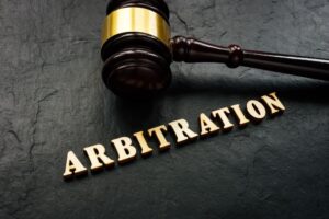 The word "Arbitration" spelled out in wooden letters, alongside a gavel in a courtroom setting. 